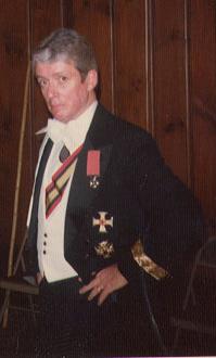Grey-haired gentleman in black tail-coat uniform with white waistcoat and white tie, sash, collar order, and other decorations.