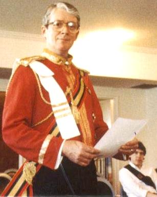 Grey=haired gentleman in glasses wearing red uniform jacket with sash, gold aiguillette, and several orders, holding a document.